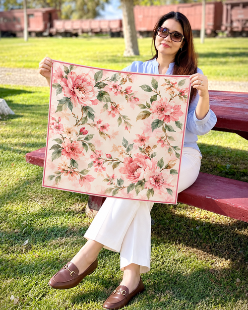 A woman sitting on a red wooden park bench holds up a square scarf with a pink and green floral print on a cream background. The setting is outdoors on a grassy lawn with trees and train cars in the background.