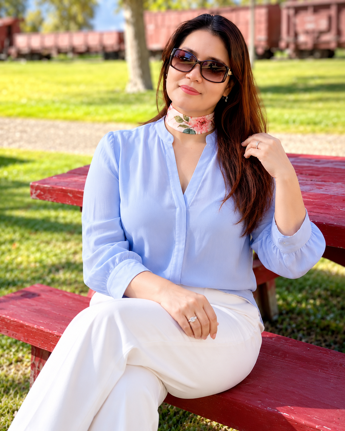 A woman wearing a light blue blouse, white trousers, and a floral choker sits on a red bench in an outdoor park setting with grass and train cars in the background.