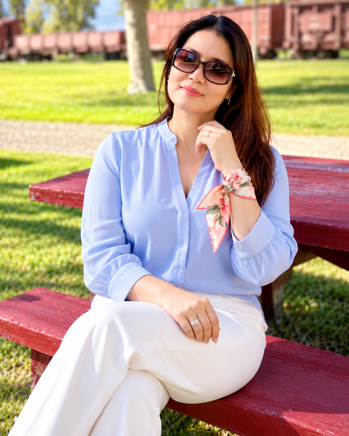 A woman wearing a light blue long-sleeve blouse and white pants sits on a red bench outdoors. She is wearing sunglasses and a pink floral wrist scarf.