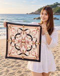 A woman standing on a sandy beach holds up a square silk scarf with an ornate baroque-style pattern in pink, black, beige, and gold.