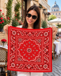 A woman wearing sunglasses holds up a red square bandana with a white and pink paisley pattern in front of an outdoor cafe setting.