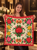A woman holds up a square silk scarf featuring a vibrant, symmetrical floral pattern with red, pink, yellow, and white flowers on a cream background with a red border. She is standing in a luxurious hotel lobby with a 'Grand Imperial Hotel' sign visible in the background.