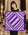 A woman in a black dress and heels holds up a square scarf with a bold geometric pattern in shades of purple, lavender, and white, set against a luxurious hotel lobby background.